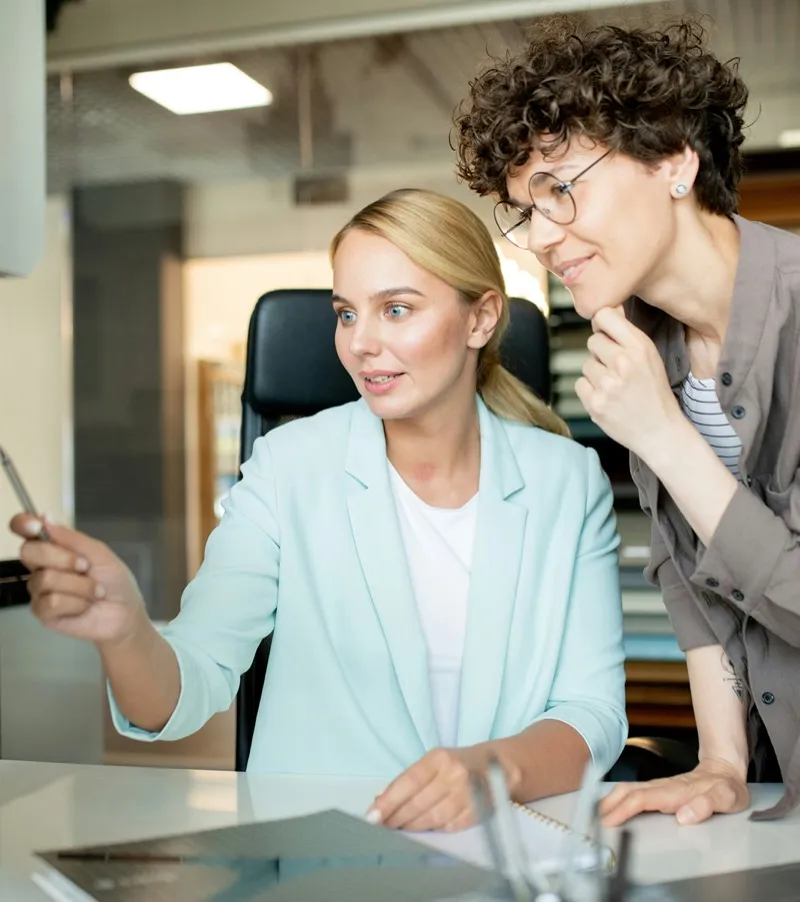 A colleague leans in to review work with a smiling woman at her desk